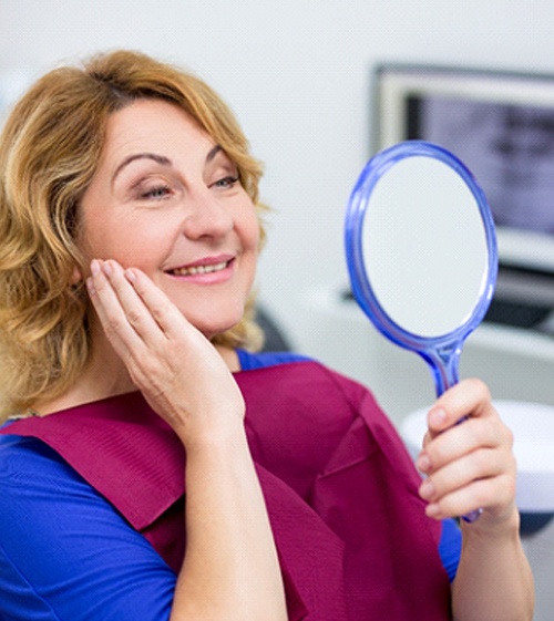 An older woman admires her new smile in the mirror after receiving dental implants