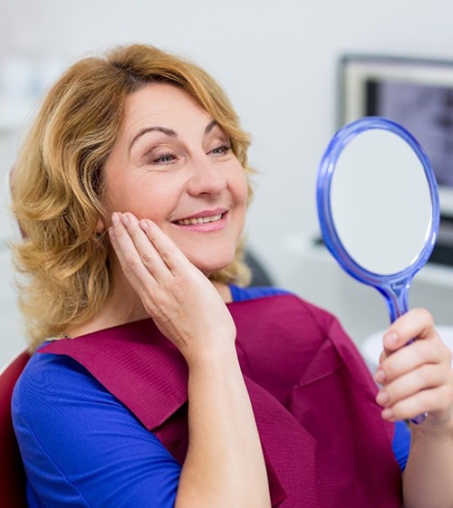 An older woman wearing a blue blouse and holding her cheek while admiring her new smile in the mirror at the dentist’s office
